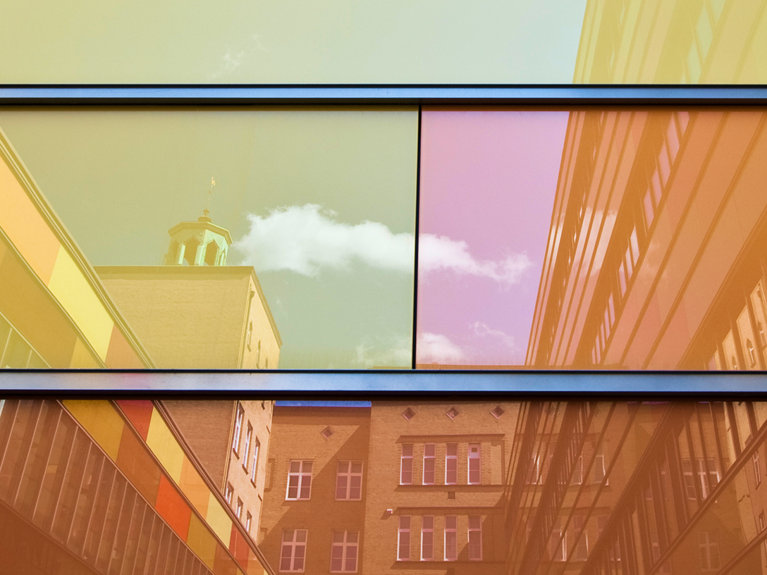 Reflektierende Fensterfassade mit bunten Farbtönen, in der sich ein Gebäude und Wolken spiegeln.