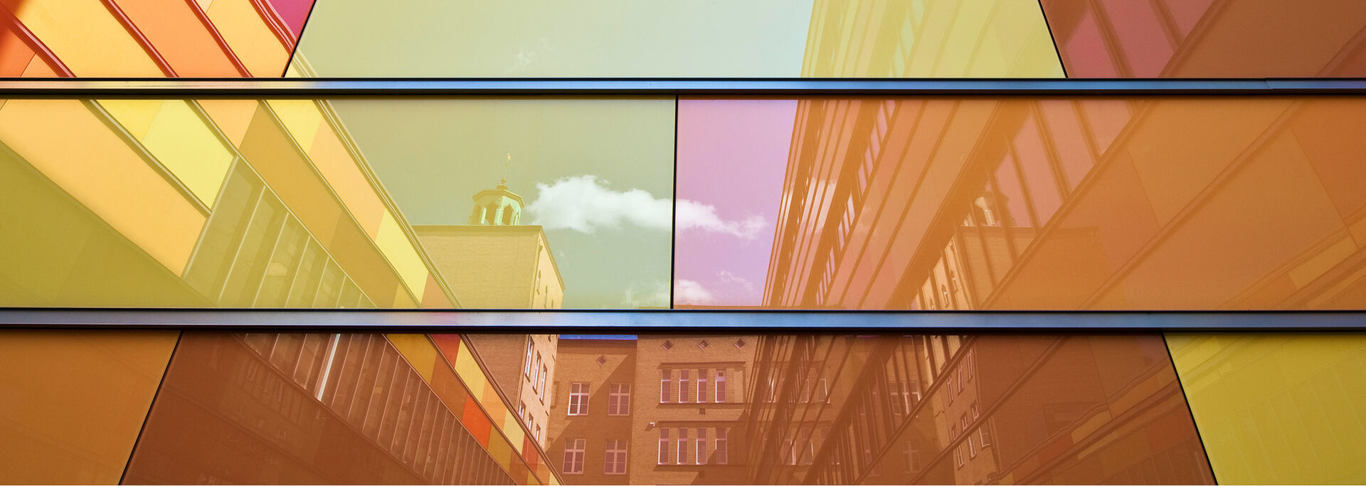 Reflektierende Fensterfassade mit bunten Farbtönen, in der sich ein Gebäude und Wolken spiegeln.