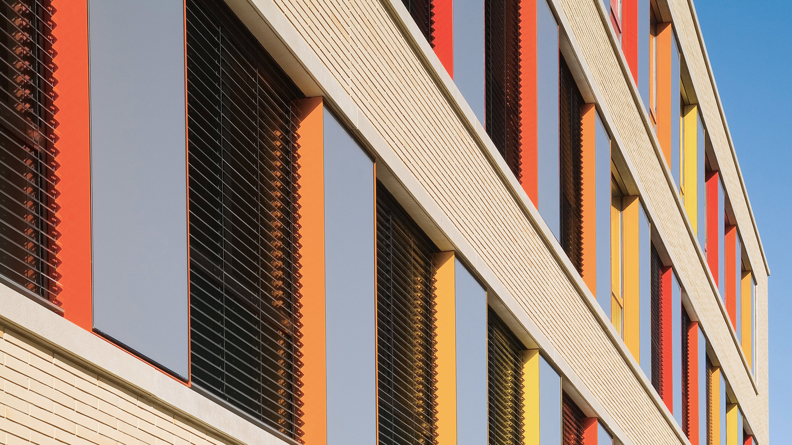 Fassade eines modernen Gebäudes mit bunten Fenstern in Rot, Gelb und Orange vor klarem Himmel.