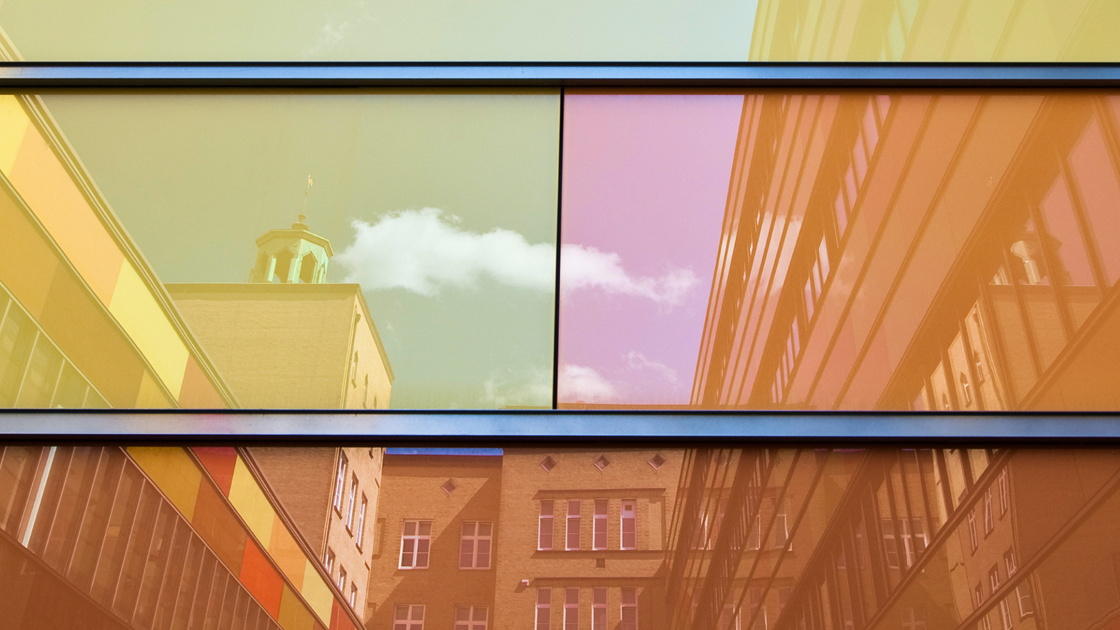 Reflektierende Fensterfassade mit bunten Farbtönen, in der sich ein Gebäude und Wolken spiegeln.