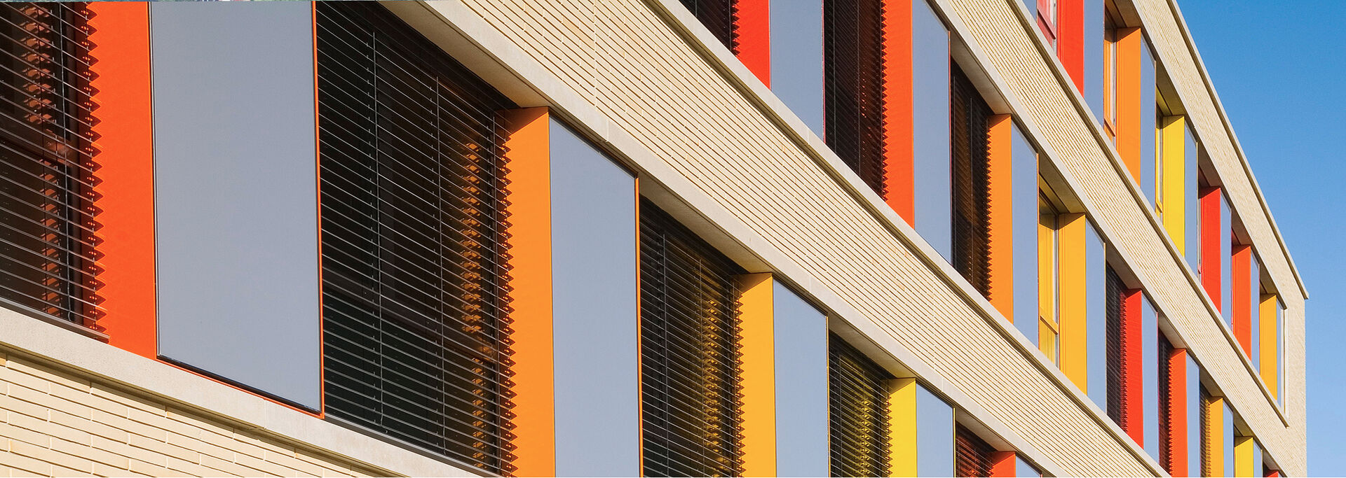 Fassade eines Gebäudes mit bunten Fenstern in Rot, Gelb und Orange vor einem blauen Himmel.