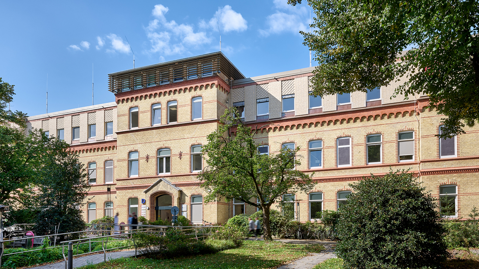 Historisches Gebäude mit vielen Fenstern und einem begrünten Vorplatz unter blauem Himmel.