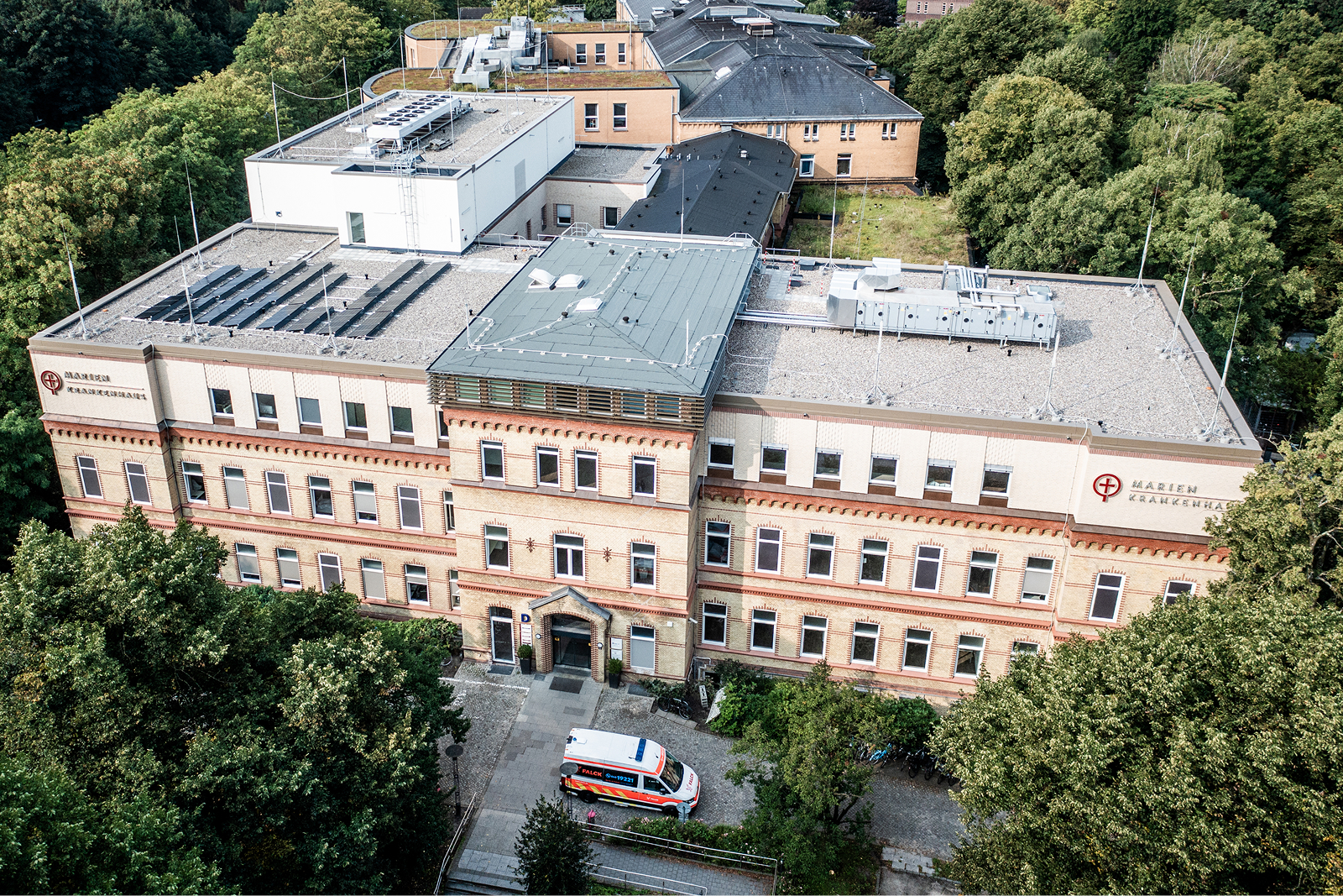 Helles, historisches Klinikgebäude aus der Vogelperspektive, umgeben von Parkanlagen und Bäumen.
