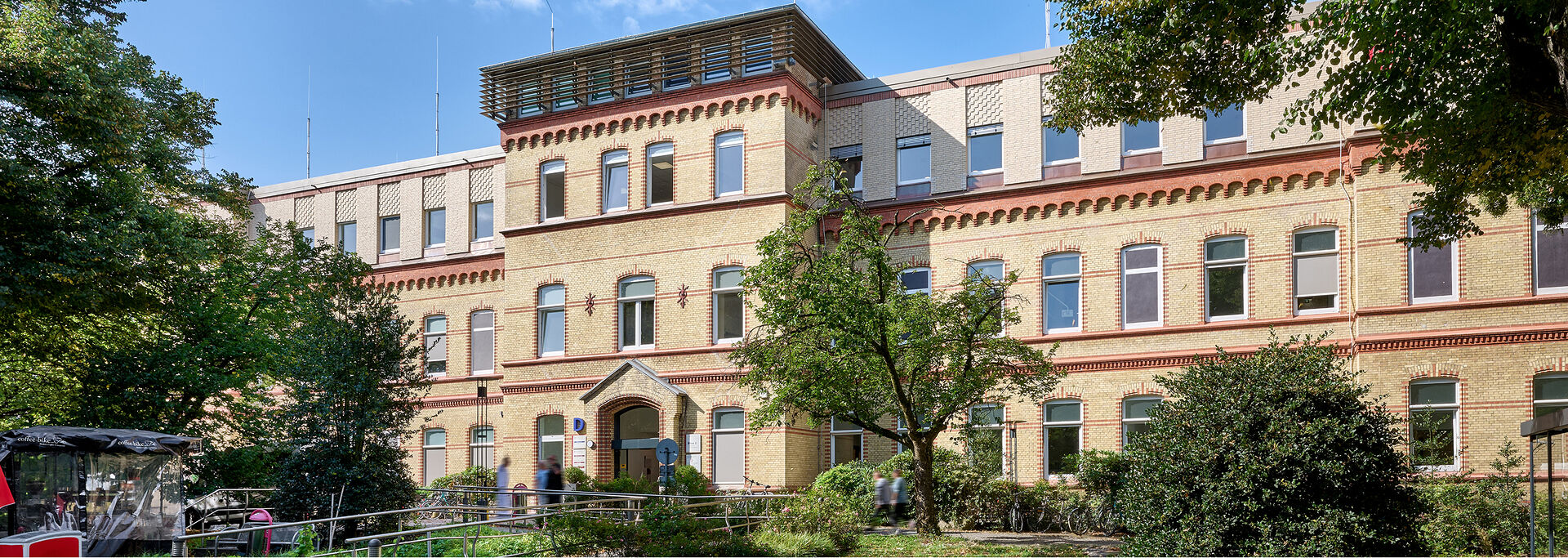 Historisches Gebäude mit mehreren Fenstern, umgeben von Bäumen und einem gepflegten Garten.