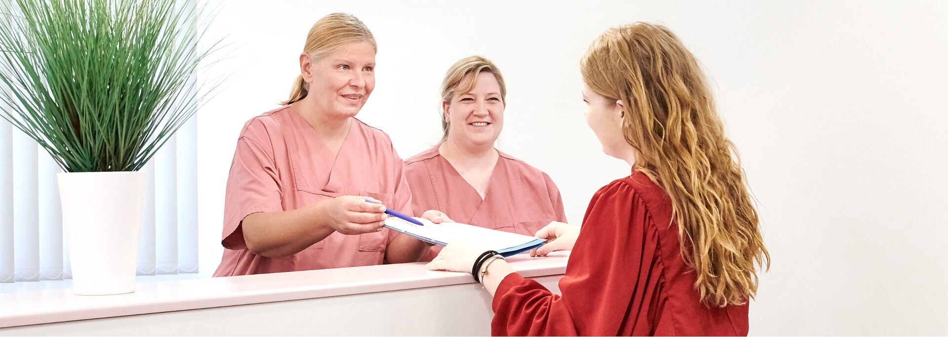 Zwei freundliche Frauen in rosafarbener Arbeitskleidung stehen hinter einem Empfangstresen und übergeben einer Patientin Dokumente.