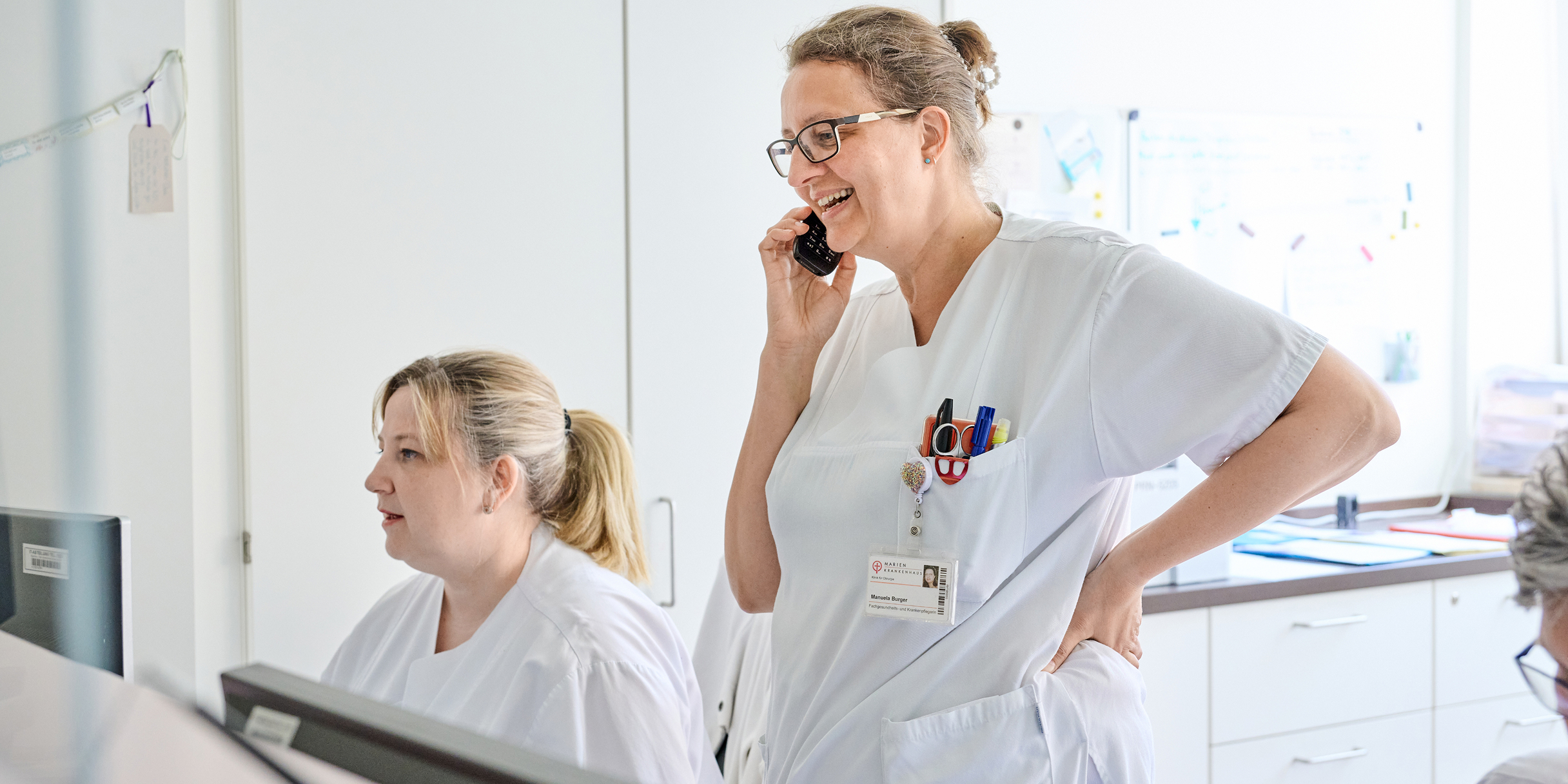 Zwei medizinische Fachkräfte in weißer Arbeitskleidung, eine telefoniert, die andere sitzt am Computer. Helle, moderne Umgebung.