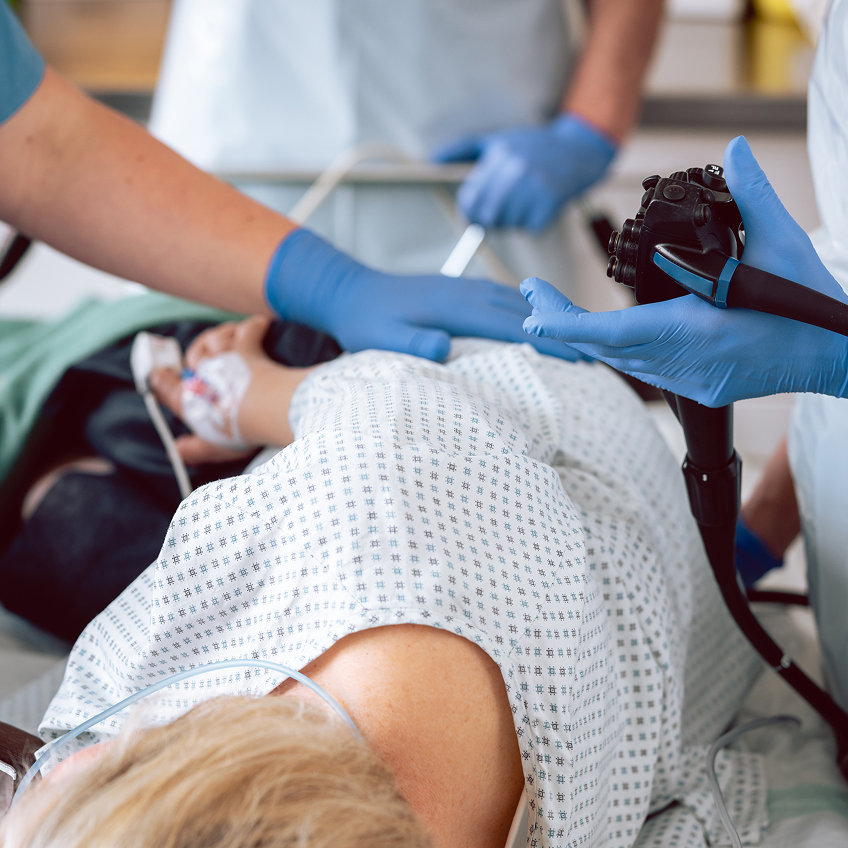 Medizinisches Personal in blauen Handschuhen führt einen Eingriff an einer Patientin in Krankenhauskleidung durch, die seitlich auf einem Behandlungstisch liegt.