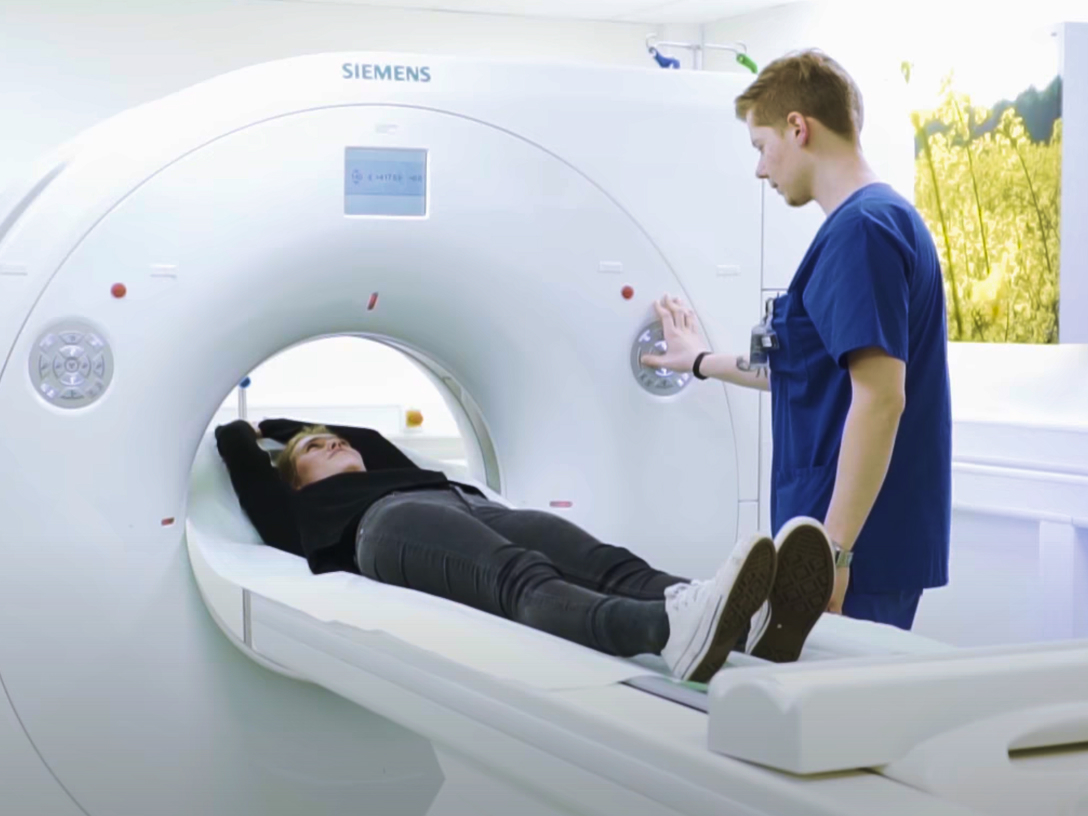 Eine Patientin liegt auf einer Liege eines CT-Gerätes, während eine medizinische Fachkraft das Gerät bedient.