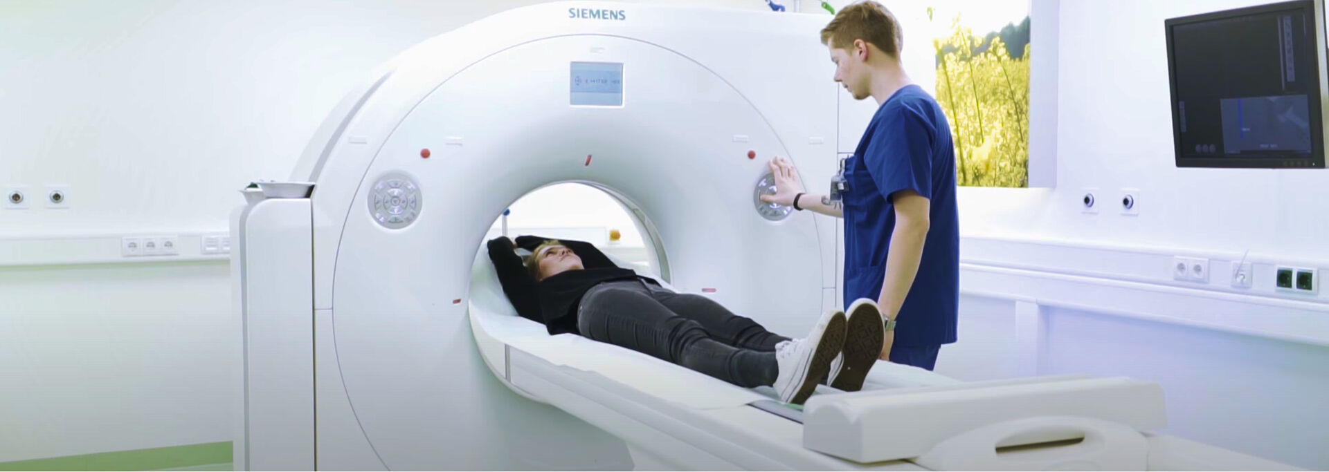 Ein Patient liegt auf einer Liege in einem CT-Scanner, während eine Fachkraft davorsteht und das Gerät bedient.