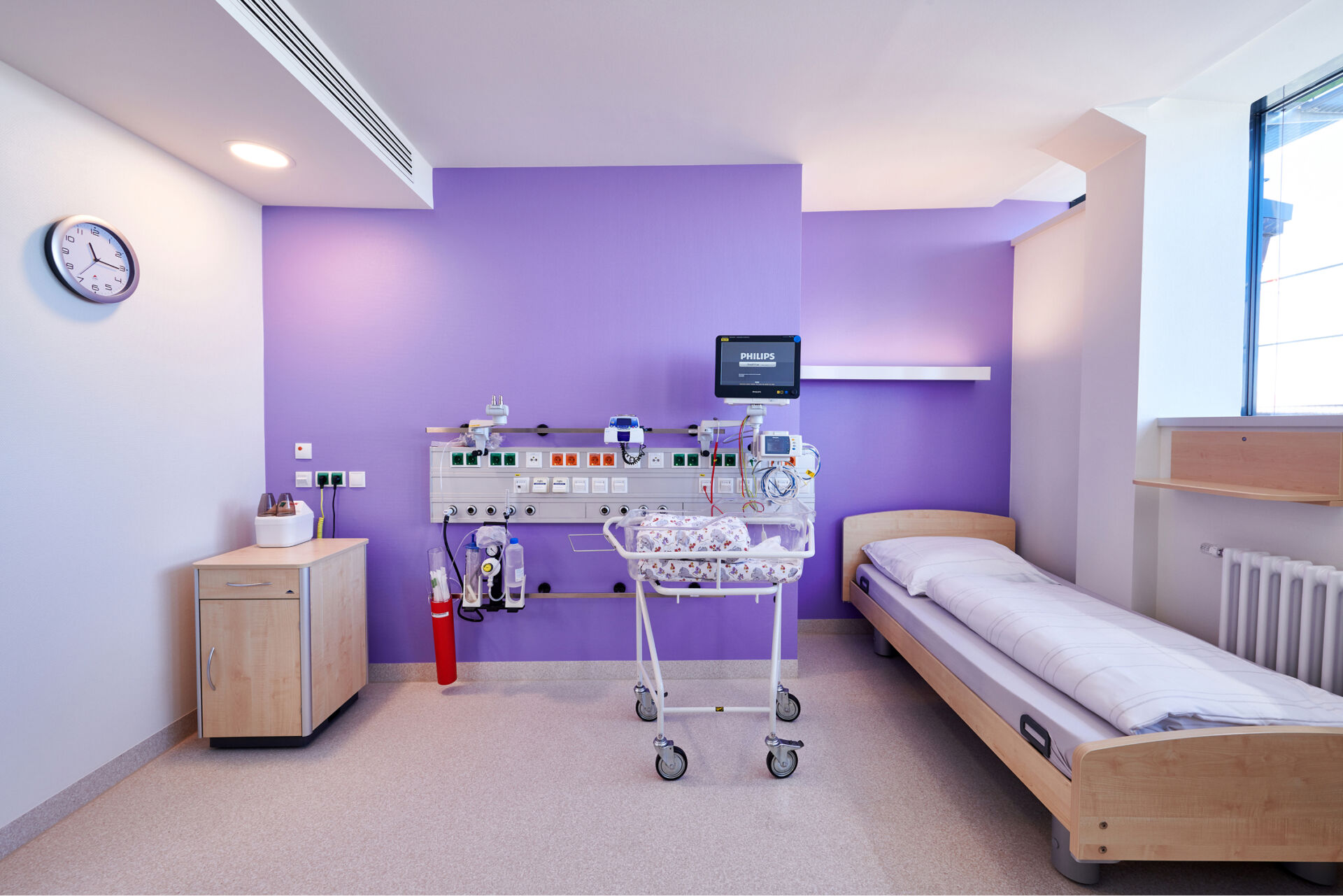 Krankenhauszimmer mit lilafarbener Wand, einem Bett, einem Babybett und medizinischen Geräten.