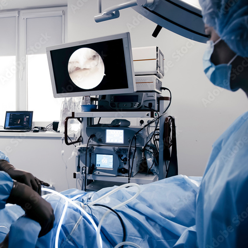 Ein OP-Team behandelt einen Patienten, der vor ihnen auf dem Tisch liegt. Im Hintergrund sieht man einen Monitor mit medizinischen Bildern.