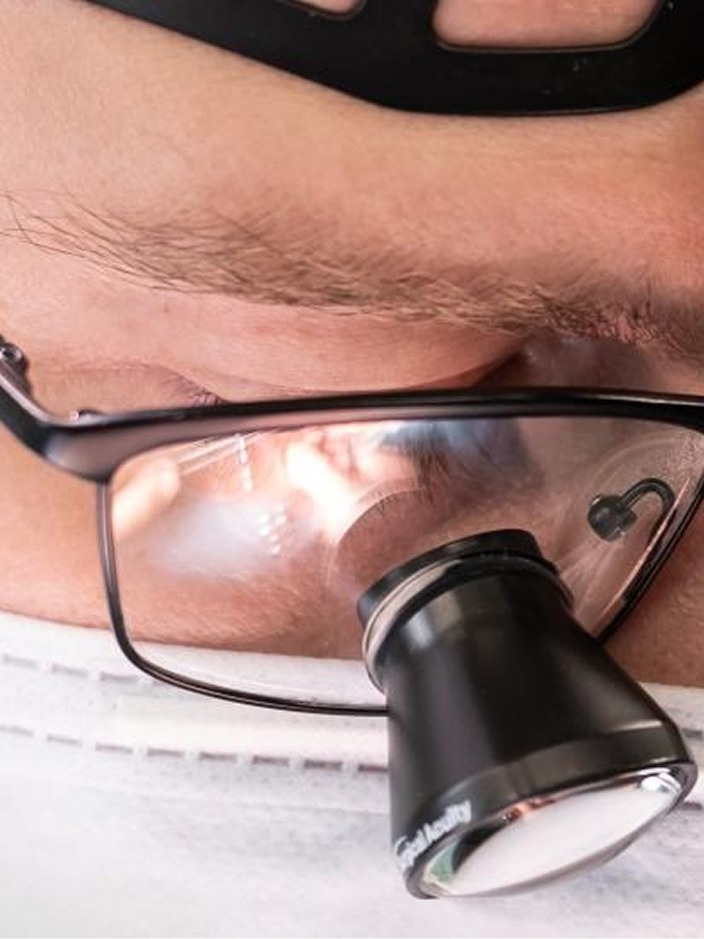 Nahaufnahme eines Gesichts mit OP-Brille und einer Lupe, die Person trägt einen weißen Mundschutz.