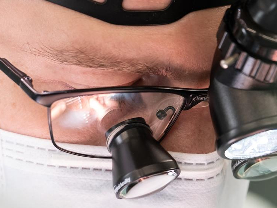 Nahaufnahme eines Gesichts mit OP-Brille und einer Lupe, die Person trägt einen weißen Mundschutz.