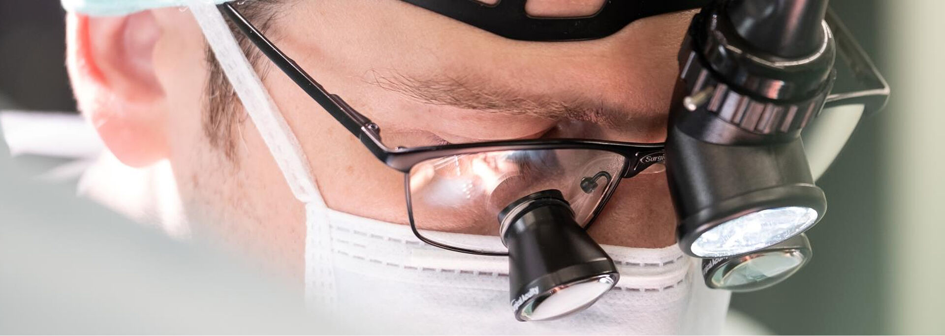 Nahaufnahme eines Gesichts mit OP-Brille und einer Lupe, die Person trägt einen weißen Mundschutz.