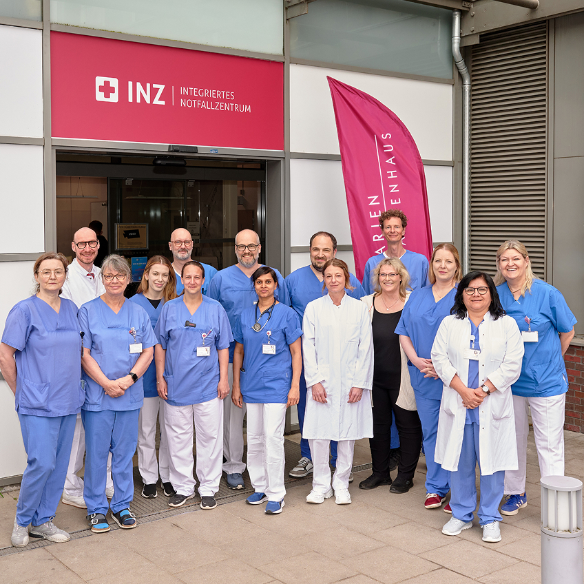 01-infos-fuer-zuweisende-02 Gruppe von medizinischen Fachkräften in blauer Arbeitskleidung vor einem Krankenhaus, im Hintergrund ein rotes Banner.