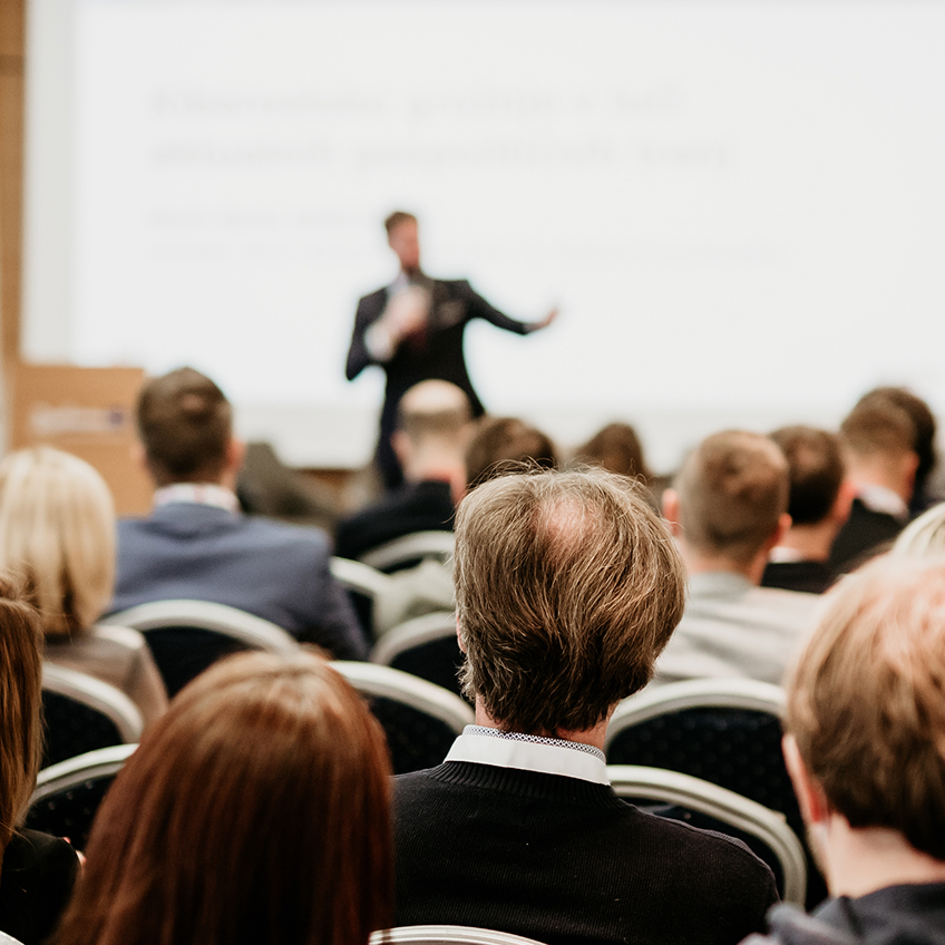 01-infos-fuer-zuweisende-03 Ein Redner steht vor einem Publikum in einem Konferenzraum, im Vordergrund sieht man die Zuschauer von hinten.