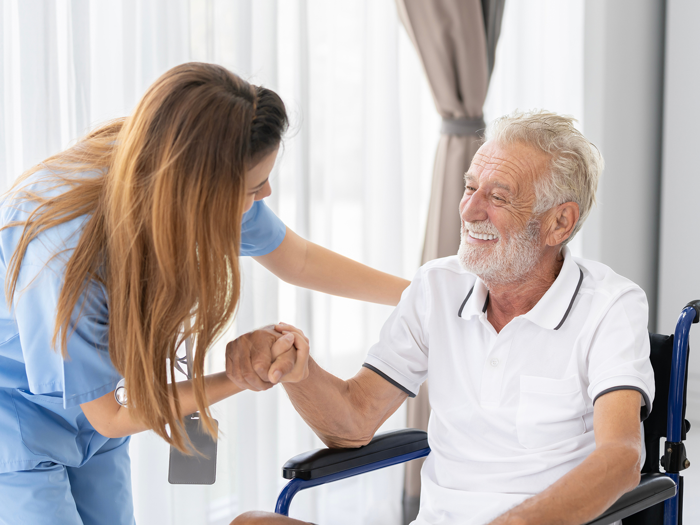 Eine weibliche Pflegekraft beugt sich lächelnd zu einem Patienten im Rollstuhl herunter und hält dabei seine Hand.