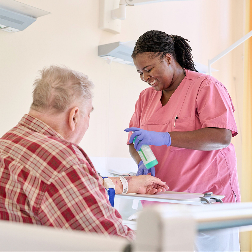 Eine Pflegekraft in rosafarbener Arbeitskleidung desinfiziert die Hand eines älteren Patienten in einem hellen Raum.