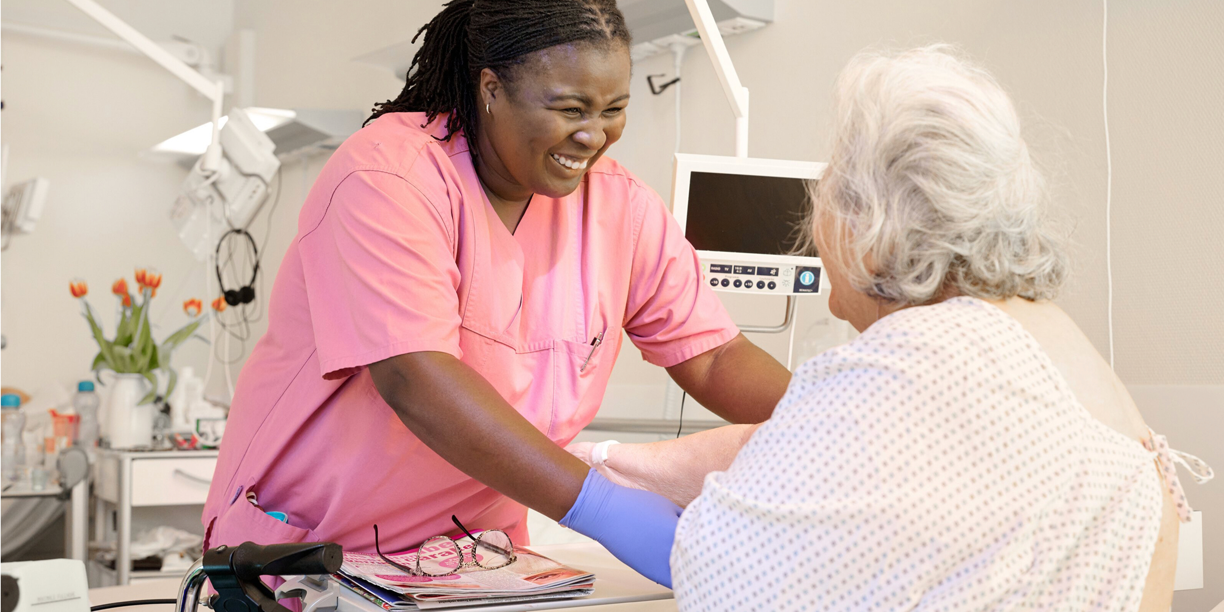 Eine lächelnde Pflegekraft in rosafarbener Arbeitskleidung interagiert freundlich mit einer älteren Patientin, die vor ihr auf dem Bett sitzt.
