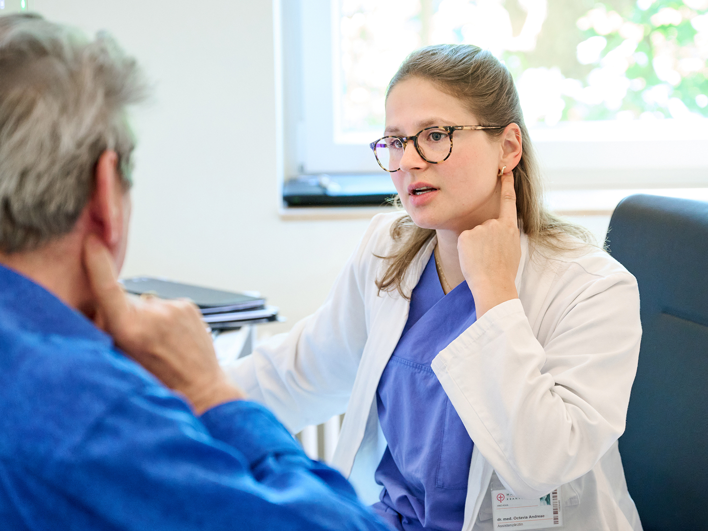 Ärztin im weißen Kittel spricht mit einem Patienten, während sie auf eine Stelle an ihrem Hals zeigt.