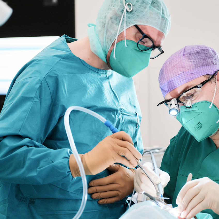 Zwei Chirurgen in OP-Kleidung und Masken führen einen Eingriff an einem Patienten durch.