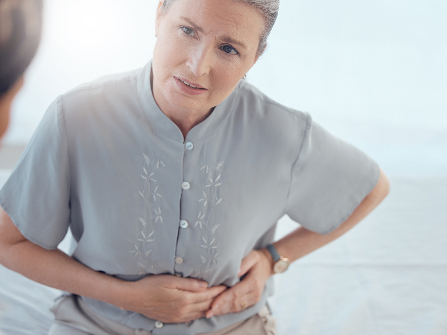 Frau mit grauem Oberteil, hält sich mit schmerzverzerrtem Gesicht den Bauch.