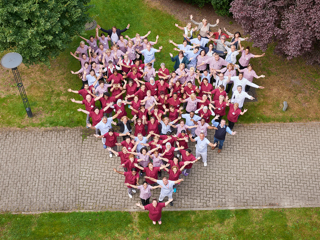 Eine große Gruppe von Menschen bildet in roter und rosafarbener Arbeitskeidung ein Herz auf einer Wiese.