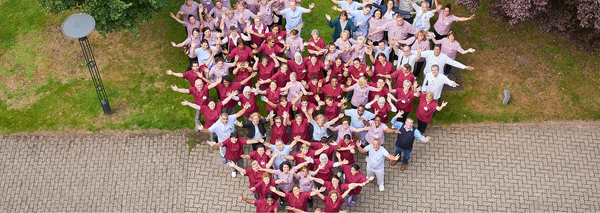 Eine große Gruppe von Menschen bildet in roter und rosafarbener Arbeitskeidung ein Herz auf einer Wiese.