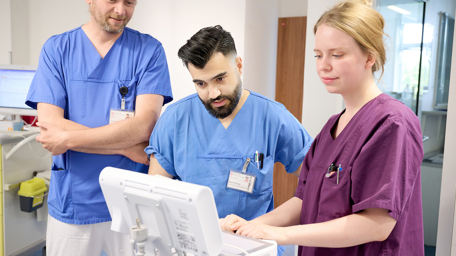 Drei medizinische Fachkräfte in blauenr und lilafarbener Arbeitskleidung betrachten gemeinsam einen Monitor.