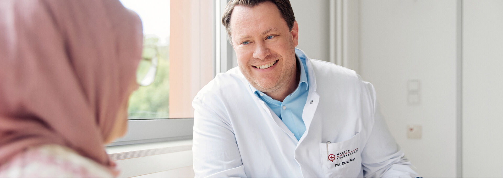 Ein lächelnder Arzt in einem weißen Kittel sitzt an einem Tisch und spricht mit einer Patientin, die ein Kopftuch trägt.
