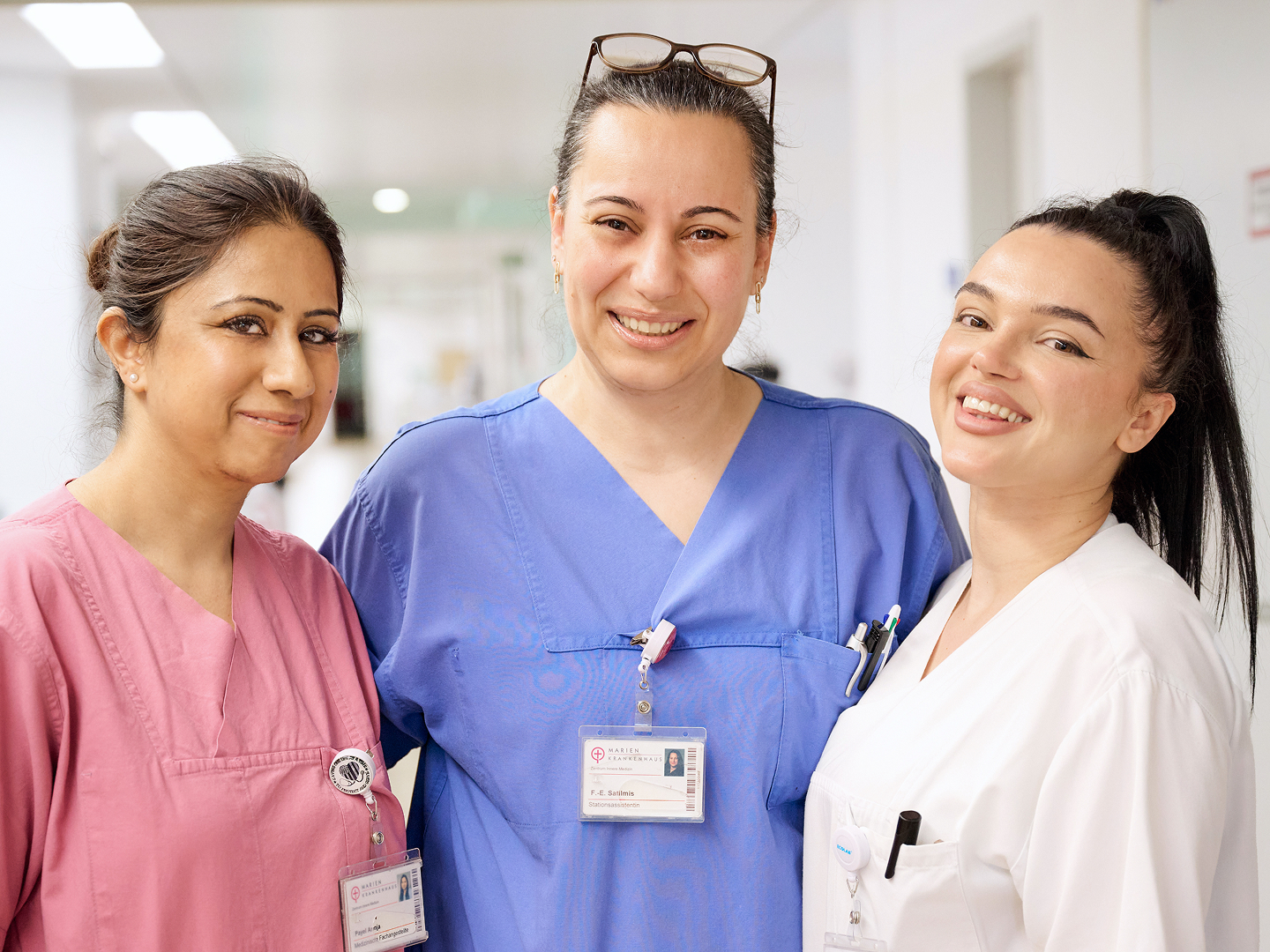 Drei Frauen in medizinischer Kleidung stehen zusammen in einem Krankenhausflur und lächeln in die Kamera.