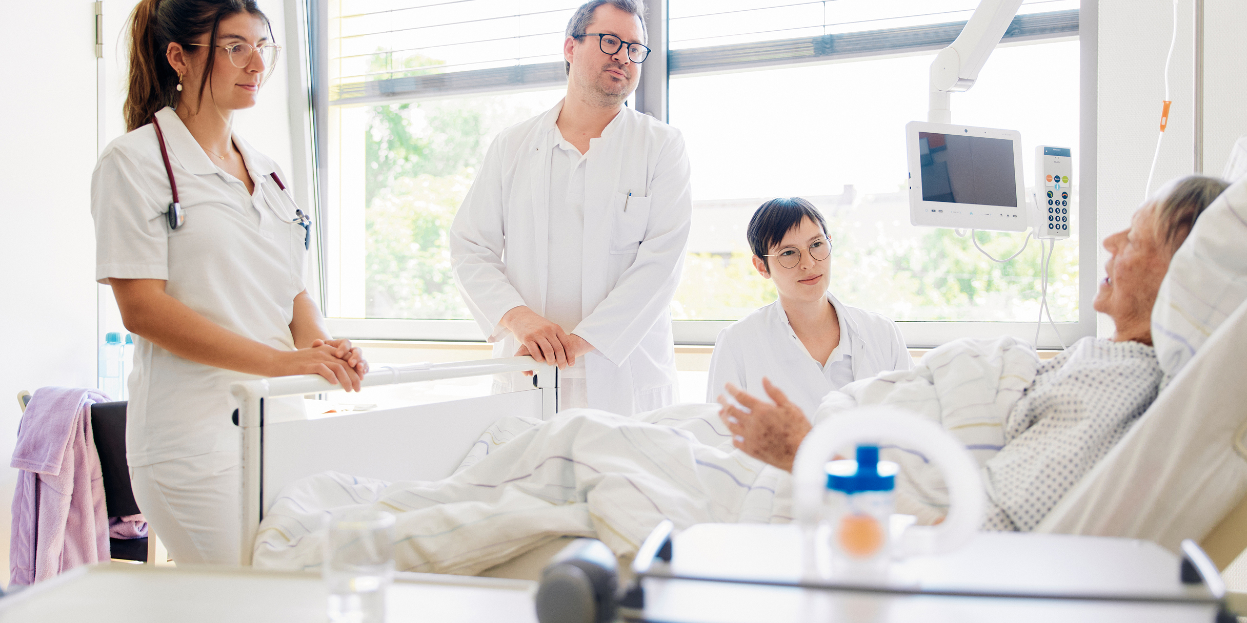 Ärzte und Pflegepersonal in weißer Kleidung stehen am Bett eines Patienten im Krankenhaus und führen ein Gespräch.