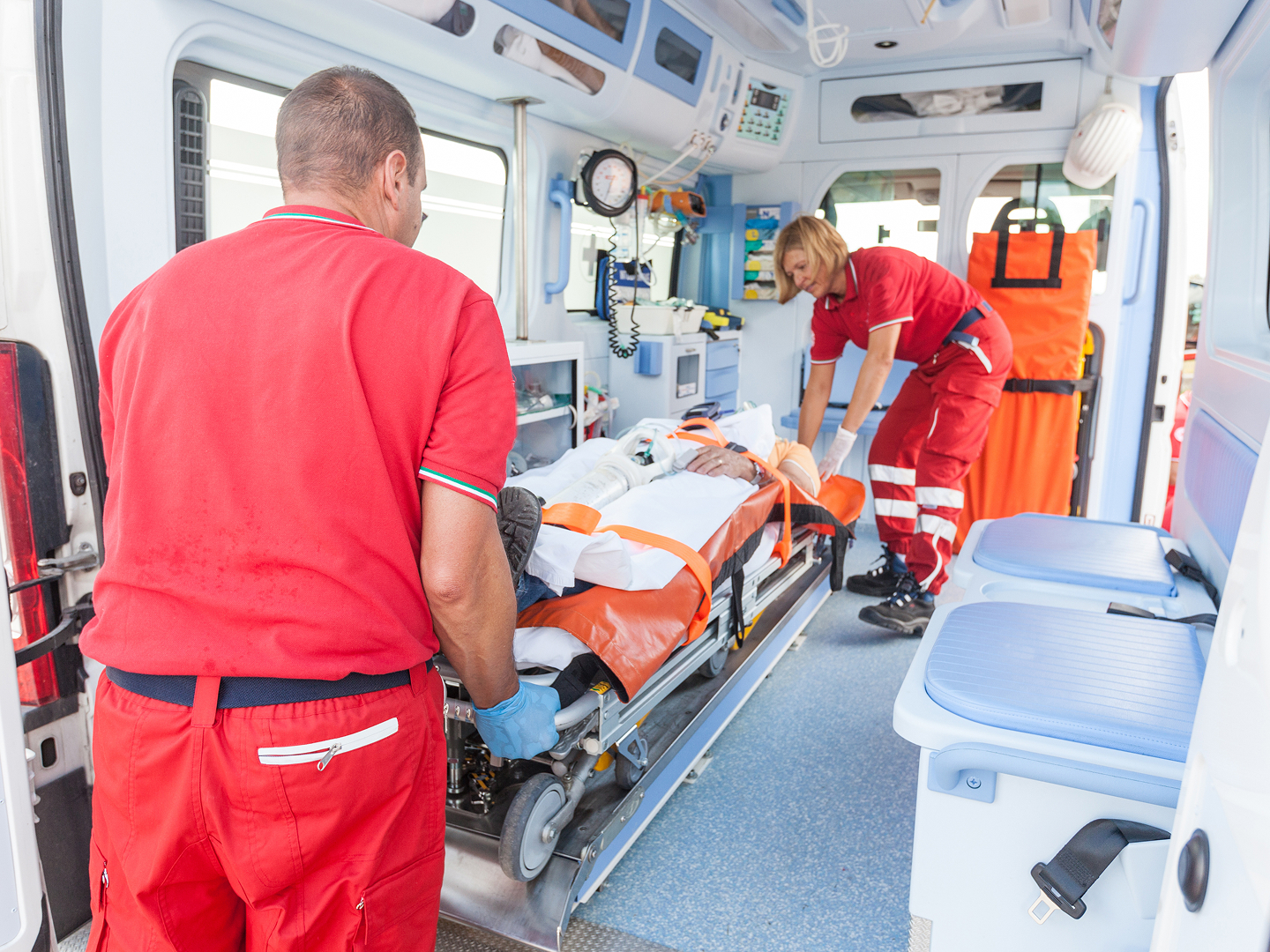 Zwei Sanitäter in roter Arbeitskleidung bringen einen Patienten auf einer Trage in einen Krankenwagen.