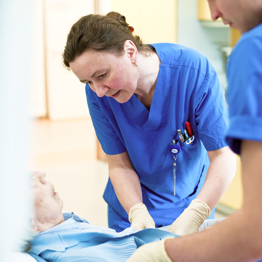 Zwei Pflegekräfte in blauer Arbeitskleidung kümmern sich um einen älteren Patienten im Bett.