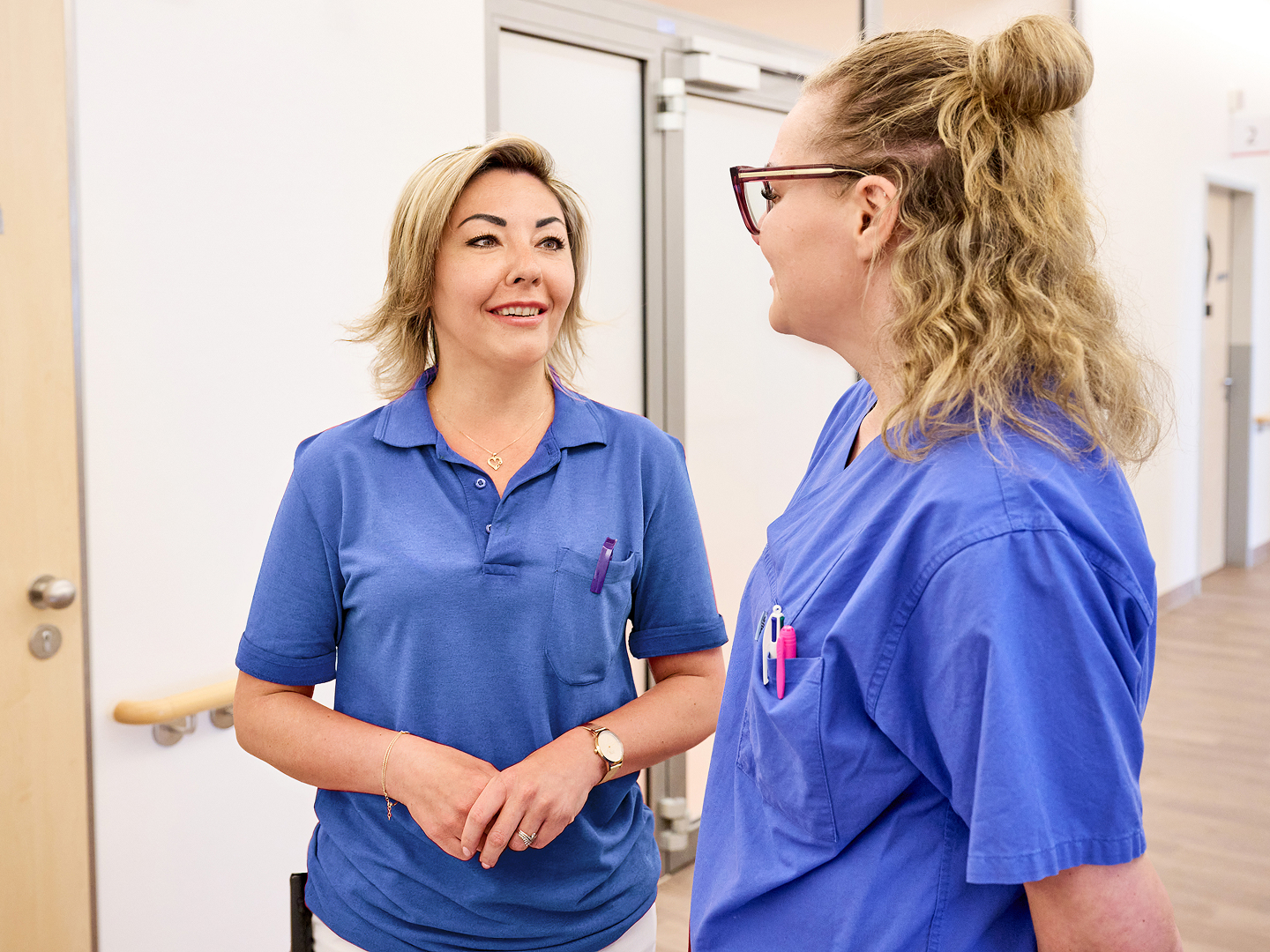 Zwei blonde Frauen in blauer Arbeitskleidung unterhalten sich im Klinikflur.