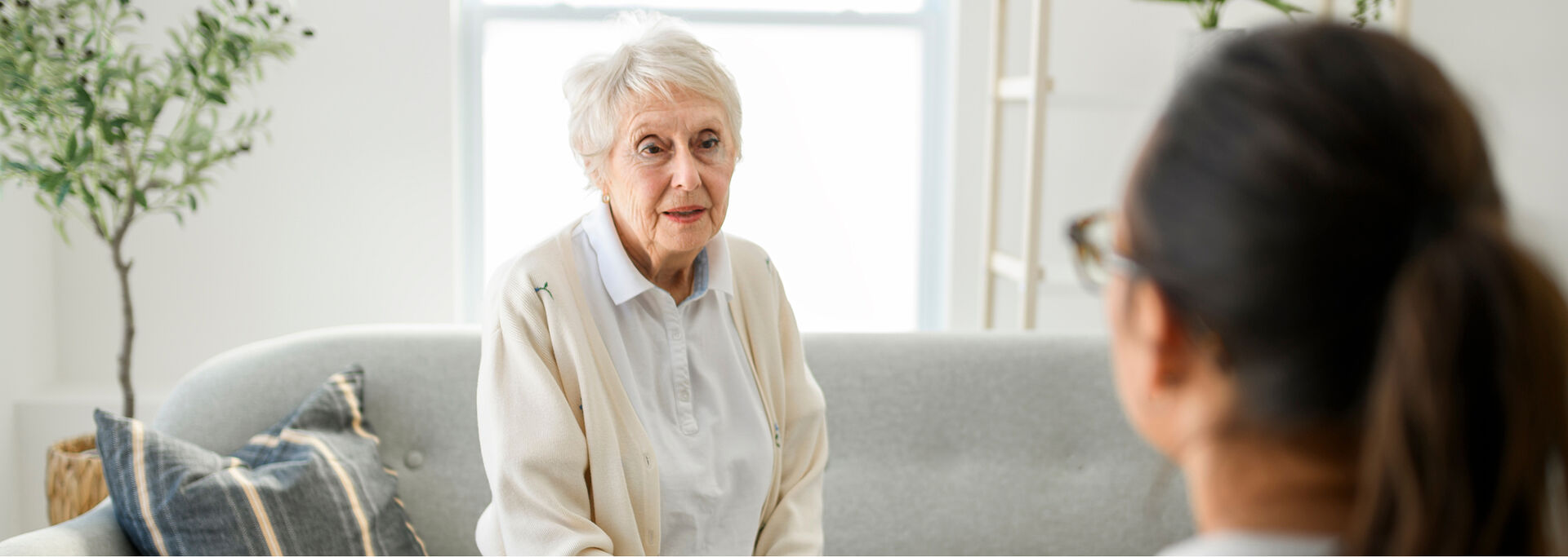 Eine ältere Patientin in heller Kleidung sitzt auf einem grauen Sofa und spricht mit einer Ärztin, die ihr gegenübersitzt.