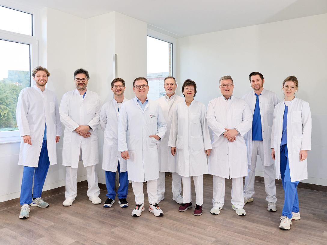 Gruppe von Ärztinnen und Ärzten in weißen Kitteln posiert in einem weißen Klinikflur für ein Foto.