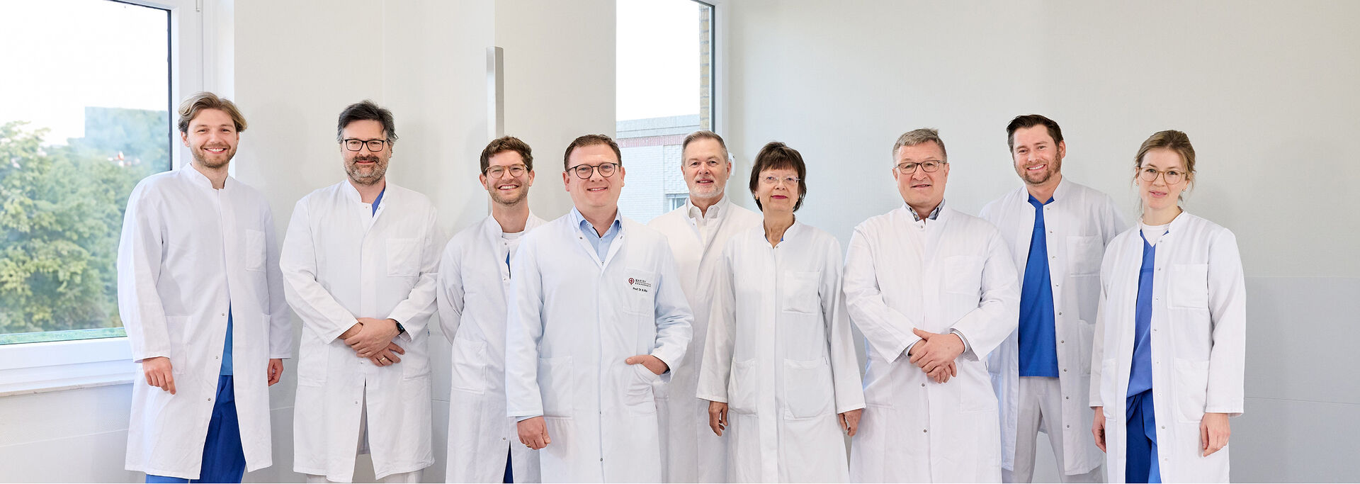 Gruppe von Ärztinnen und Ärzten in weißen Kitteln posiert in einem weißen Klinikflur für ein Foto.
