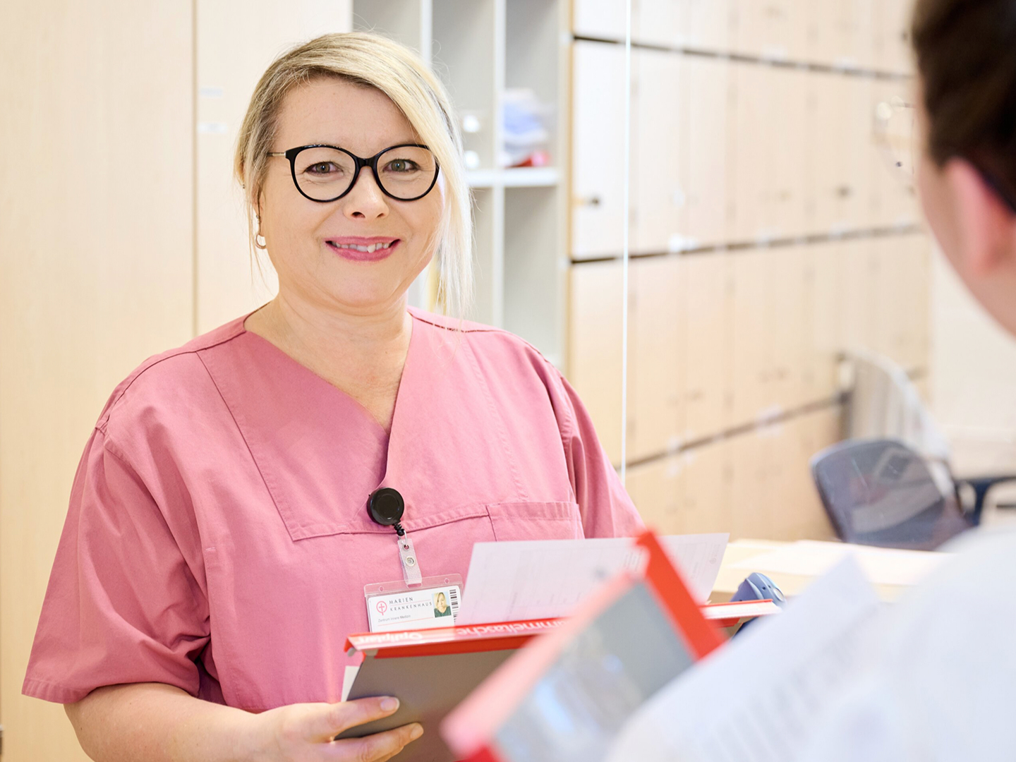 Lächelnde Frau in rosa Arbeitskleidung hält Unterlagen in der Hand, sie steht in einem hellen Raum vor einem hohen Schrank.
