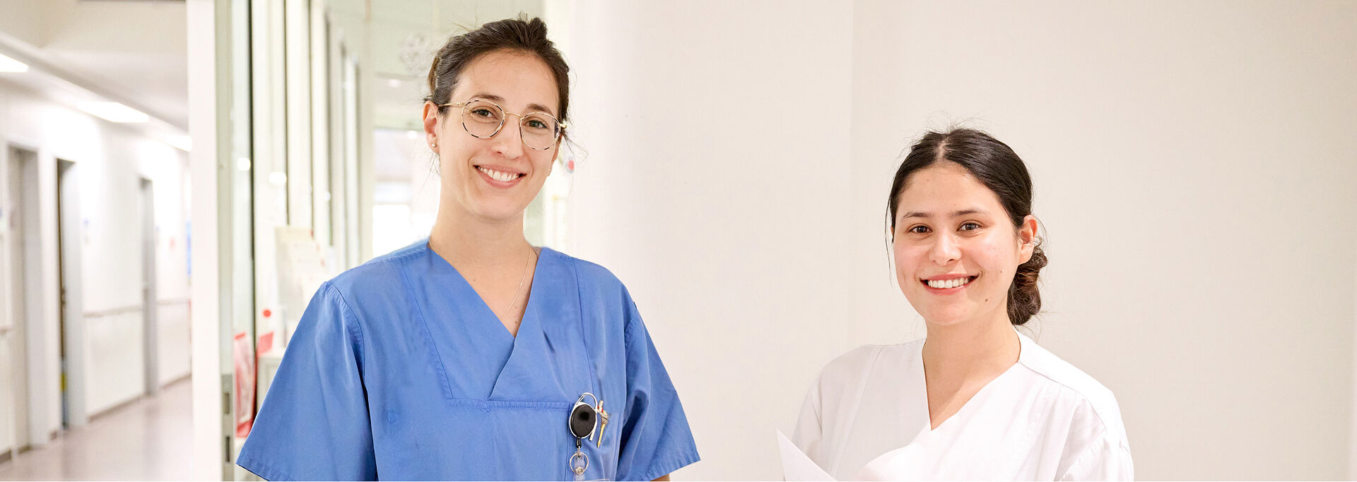 Zwei lächelnde Pflegekräfte stehen in einem Krankenhausflur, eine in blauer und die andere in weißer Arbeitskleidung.