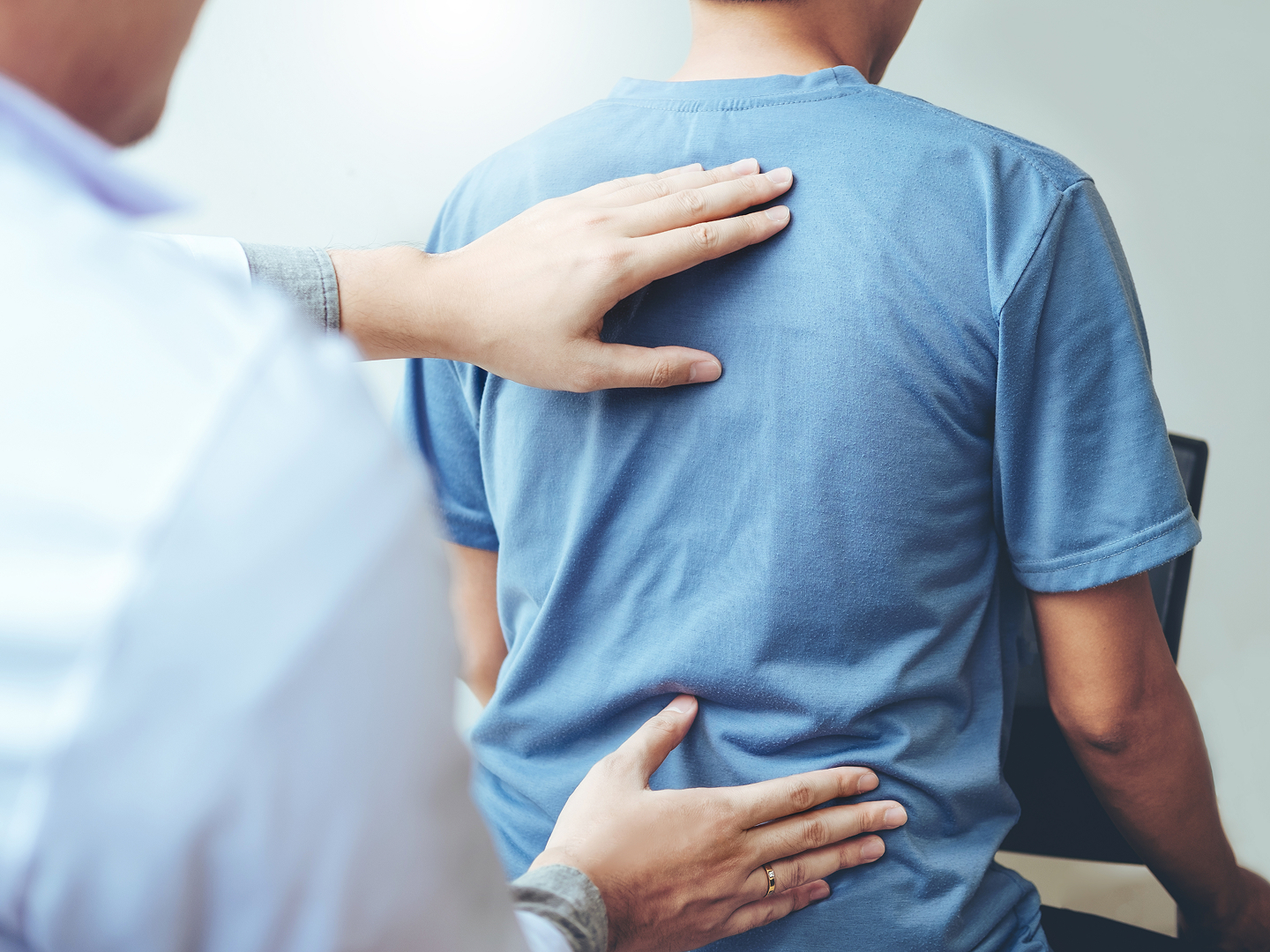 Ein Arzt untersucht den Rücken eines Patienten, der ein blaues T-Shirt trägt und auf einem Stuhl sitzt.