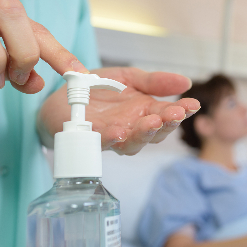 Eine Person drückt auf einen Spender mit Desinfektionsmittel, während im Hintergrund eine Person im Krankenhausbett liegt.