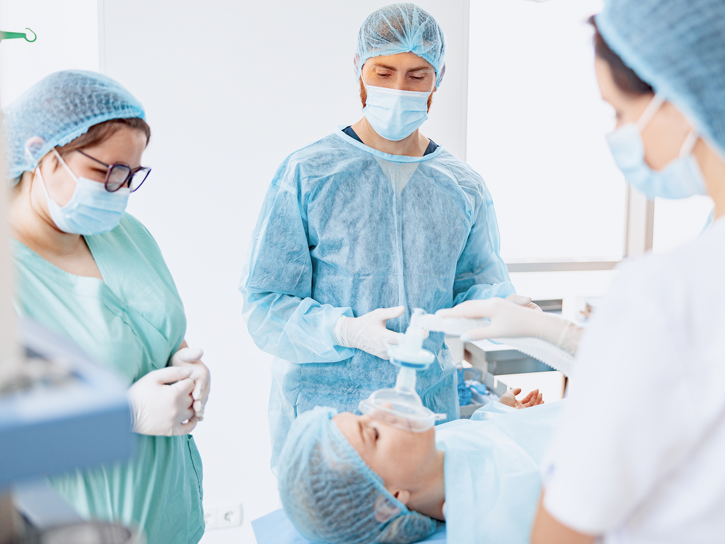 Drei medizinische Fachkräfte in Schutzkleidung und Masken stehen um einen Patienten, eine von ihnen legt ihr eine Atemmaske an.