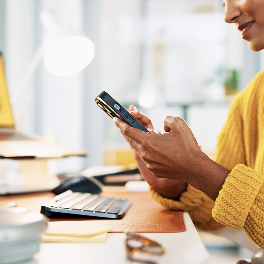 Eine Person in einem gelben Pullover hält ein Smartphone in den Händen, während sie an einem Schreibtisch mit Tastatur und Lampe sitzt.