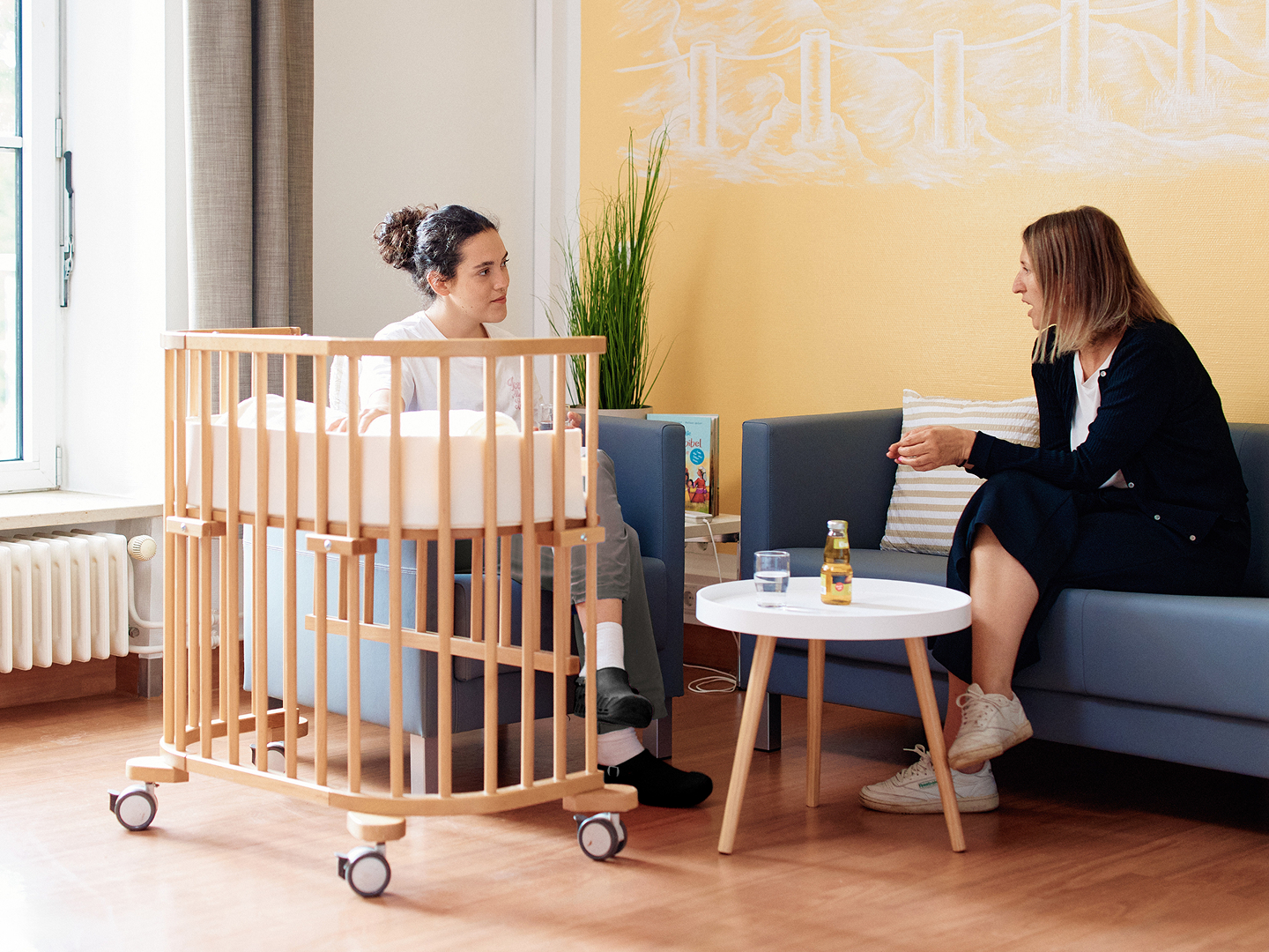 Zwei Frauen sitzen in einem modernen Raum neben einem Babybett und einem Tisch, eine spricht, während die andere zuhört.