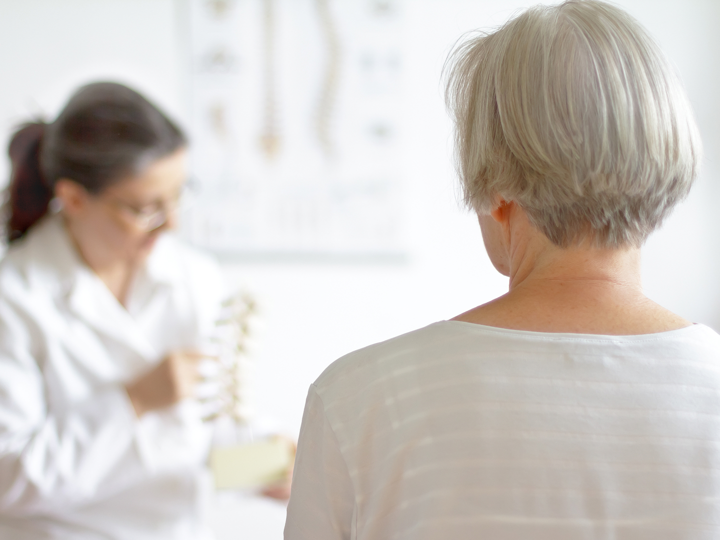 Man sieht eine ältere Frau von hinten, sie schaut zu einer Ärztin, die etwas anhand eines Wirbelsäulenmodells erklärt.