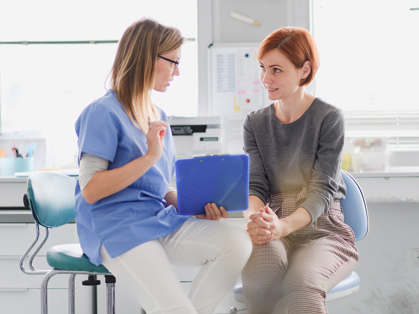 Ärztin im blauen Kittel spricht mit einer Patientin, beide sitzen sich in einem hellen Raum gegenüber.