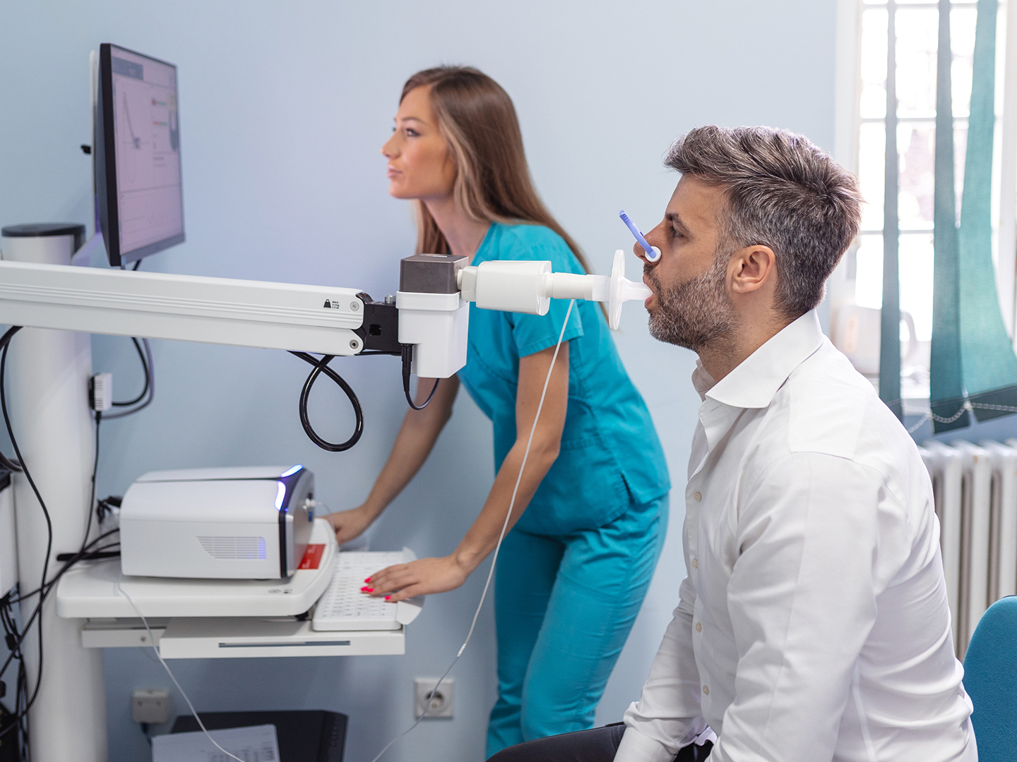 Eine Frau in blauer Arbeitskleidung prüft einen Monitor, während ein Patient ein medizinisches Gerät benutzt.