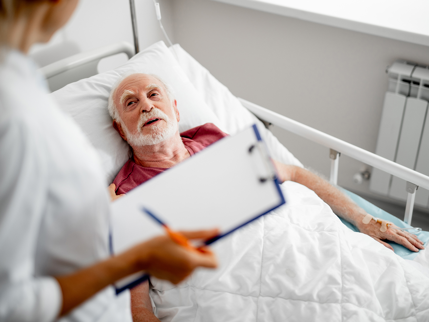 Ein älterer Mann liegt im Krankenhausbett und spricht mit einer Ärztin, die ein Klemmbrett hält und sich Notizen macht.