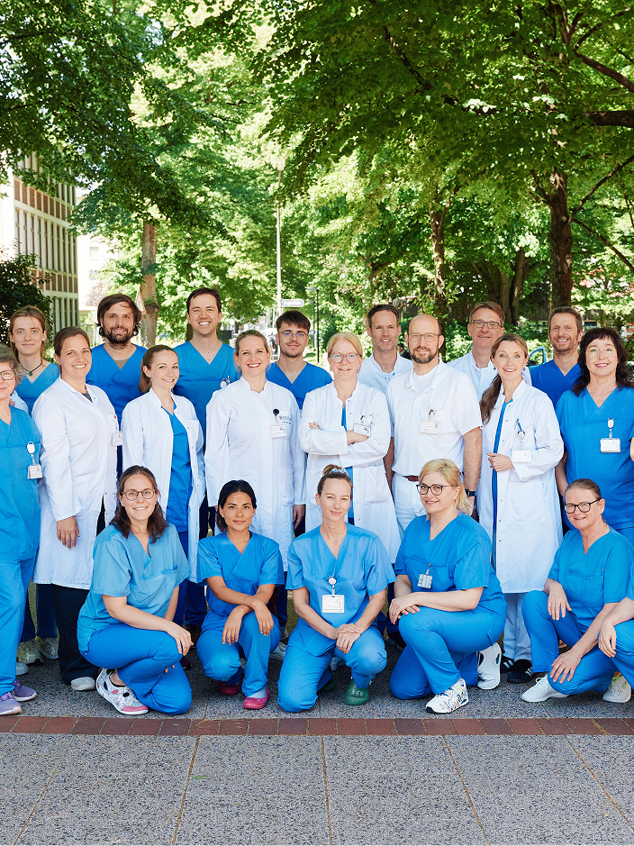 Gruppe von medizinischem Personal in blauen und weißen Kitteln steht m Freien vor Bäumen.
